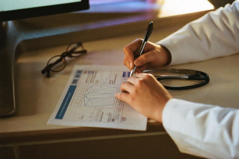 A physician's hands annotate a printed chart with a pen beside a stethoscope in warm window light.